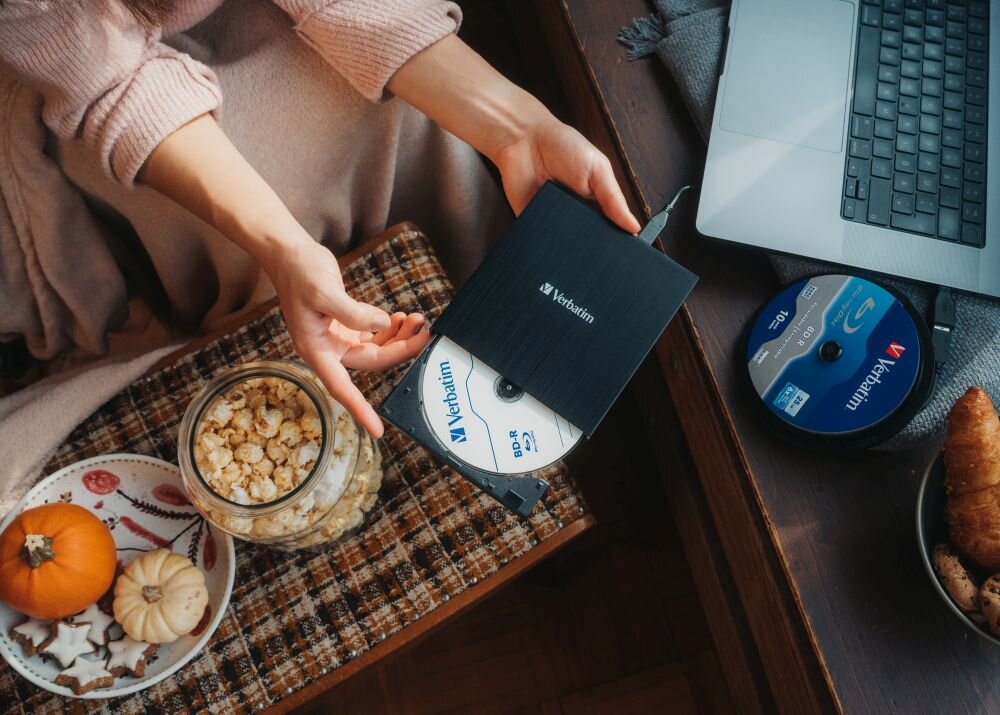 Nagrywarka Blu-ray VERBATIM Osoba wkłada płytę do czarnego napędu podłączonego do laptopa stojącego na stole, obok znajduje się słoik z popcornem, talerz z ciasteczkami i dyniami oraz stos płyt, opis