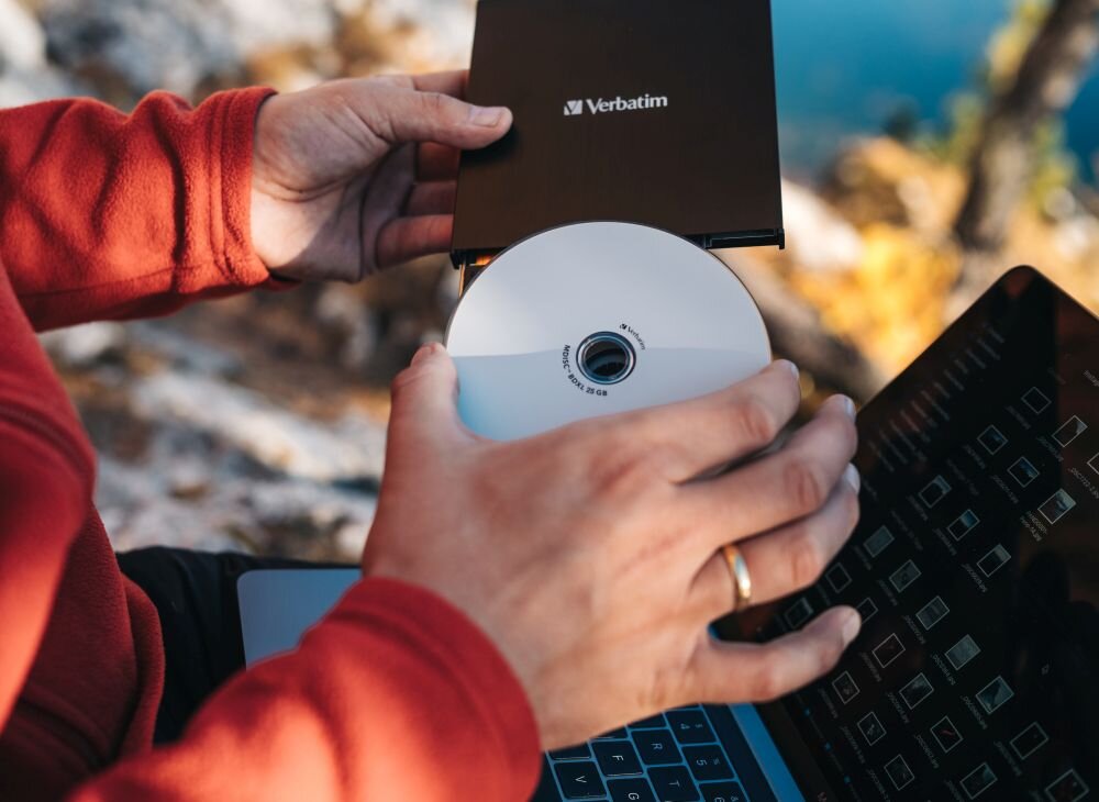 Nagrywarka Blu-ray VERBATIM Osoba w czerwonej bluzie trzyma biały krążek i umieszcza go w czarnym napędzie podłączonym do laptopa, na ekranie widoczne są miniatury plików Ultracienka nagrywarka Blu-ray – lekka i przenośna