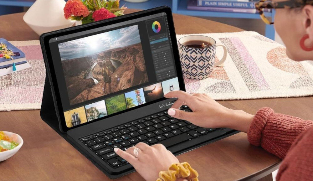 Osoba używa tabletu z klawiaturą, na ekranie widoczny jest program do edycji zdjęć. W tle znajduje się filiżanka z napojem i wazon z kwiatami.