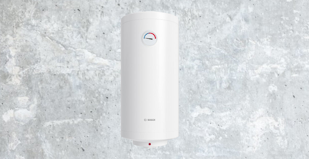 Na tle szarej, betonowej ściany znajduje się biały bojler elektryczny z logo BOSCH. Na bojlerze widoczny jest wskaźnik temperatury z kolorami niebieskim i czerwonym.