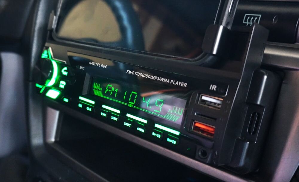 Radio samochodowe z wyświetlaczem pokazującym częstotliwość FM 107.9, z zielonym podświetleniem przycisków i portami USB. Widoczny napis: \'NAVITEL RD6 FM/BT/USB/SD/MP3/WMA PLAYER\'.