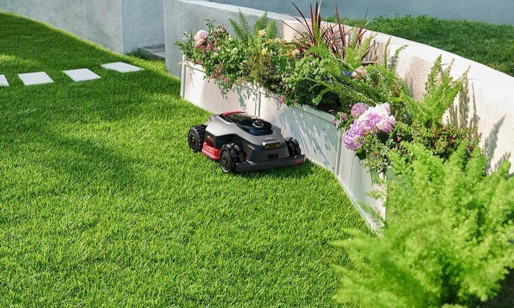 Robot koszący porusza się po zielonym trawniku obok betonowej ściany z kwiatami i roślinami. W tle widoczne są białe płytki chodnikowe.