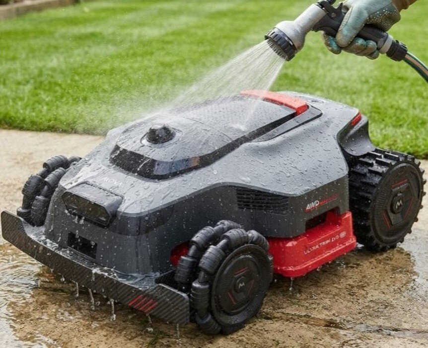 Robot koszący jest myty wodą z węża ogrodowego na betonowej powierzchni. Na urządzeniu widoczne są napisy: \'AWD\' i \'ULTRA TRIM DQ\'.