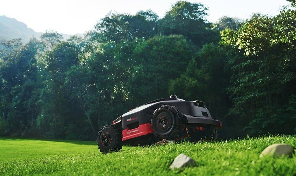 Robot koszący na trawie, otoczony zielonymi drzewami w tle. Urządzenie ma czarną i czerwoną obudowę oraz widoczne duże koła.