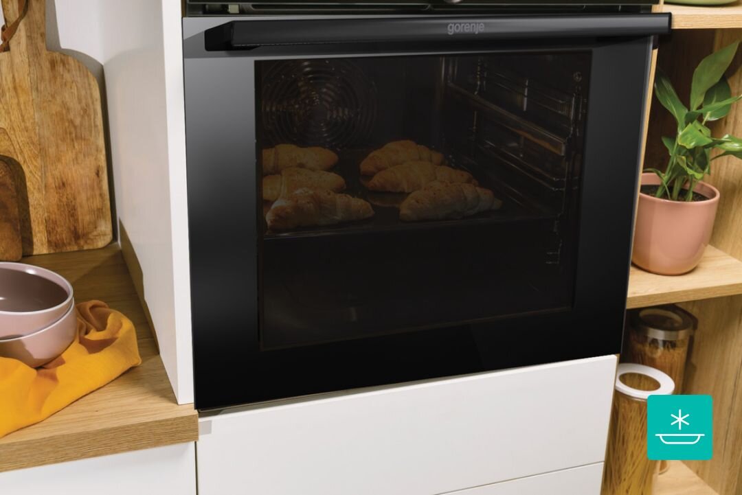 Czarny piekarnik z logo \'gorenje\' z widocznymi pieczonymi rogalikami wewnątrz. Obok piekarnika znajdują się drewniana deska, miska i roślina w doniczce.