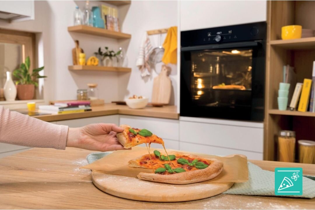 W kuchni na drewnianym blacie leży pizza z bazylią, z której ktoś wyciąga kawałek. W tle widać czarny piekarnik z włączonym światłem wewnętrznym.