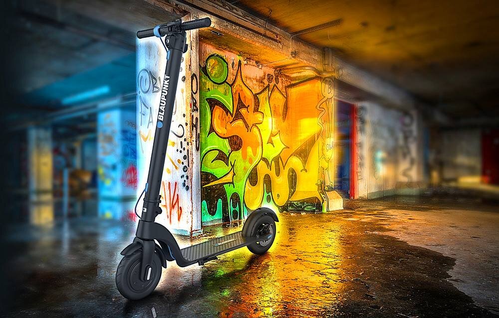 Czarna hulajnoga elektryczna z logo BLAUPUNKT stoi w kolorowym, graffiti ozdobionym pomieszczeniu. Tło jest rozmyte, z widocznymi jasnymi i ciemnymi kolorami.