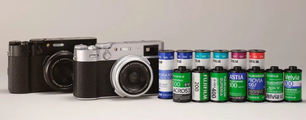 Na obrazie znajdują się dwa aparaty fotograficzne, jeden czarny i jeden srebrny, oraz szereg kolorowych rolek filmów fotograficznych z widocznymi napisami takimi jak \'FUJIFILM\', \'PROVIA 100F\', \'VELVIA 100\'.