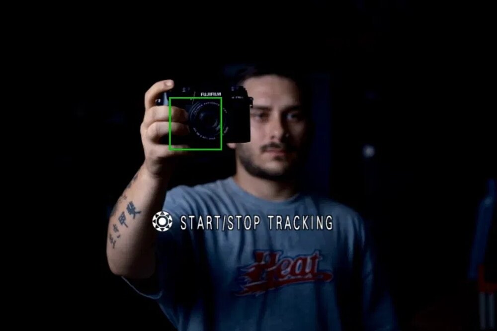 Osoba trzyma aparat marki FUJIFILM, na ekranie widoczny jest zielony prostokąt wokół obiektywu. Na obrazie znajduje się tekst: \'START/STOP TRACKING\'.