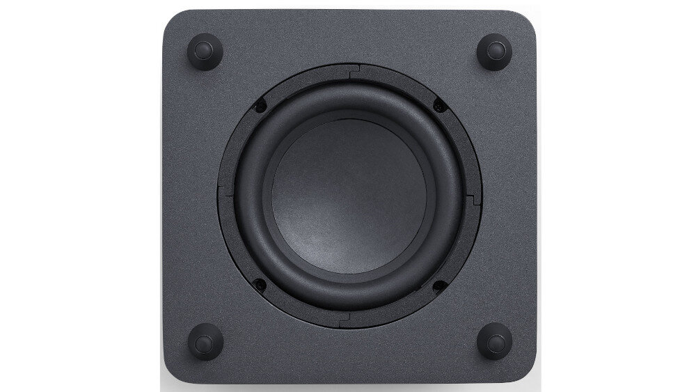 Obraz przedstawia czarny subwoofer z widocznym głośnikiem na środku i czterema gumowymi nóżkami w rogach. Powierzchnia subwoofera jest matowa.