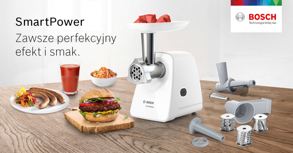 Na drewnianym stole znajduje się maszynka do mięsa z logo BOSCH, obok której leżą różne akcesoria. W tle widoczne są potrawy oraz napis \'SmartPower Zawsze perfekcyjny efekt i smak.\'