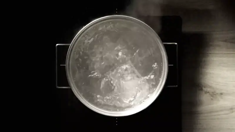 Na obrazie widoczny jest garnek z gotującą się wodą na płycie indukcyjnej. Płyta ma ciemną powierzchnię i widoczne są na niej linie oznaczające pola grzewcze.