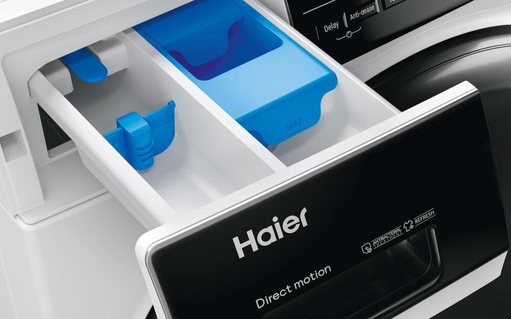 Zbliżenie na szufladę na detergenty pralko-suszarki z widocznymi niebieskimi przegródkami. Na froncie szuflady znajduje się logo \'Haier\' oraz napisy \'Direct motion\'.