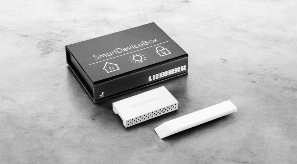 Na obrazie znajduje się czarne pudełko z napisem \'SmartDeviceBox\' oraz logo \'LIEBHERR\'. Obok pudełka leży biały, prostokątny przedmiot z perforacjami.