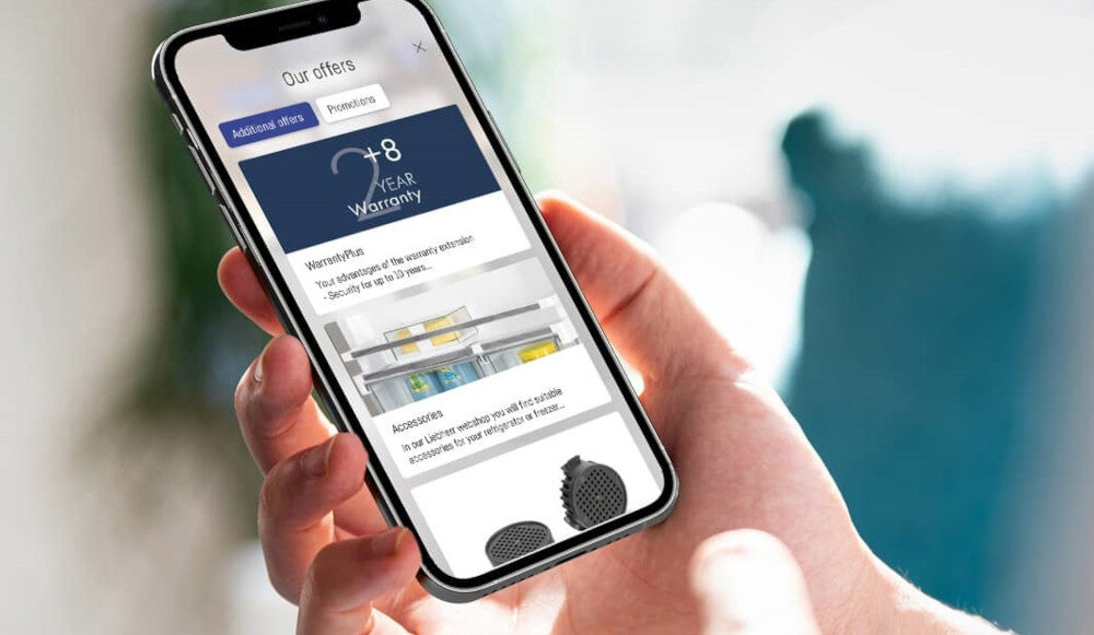 Osoba trzyma smartfon, na ekranie którego widoczna jest oferta z tekstem \'Our offers\', \'8 YEAR Warranty\', \'WarrantyPlus\', oraz zdjęcie wnętrza lodówki z akcesoriami.