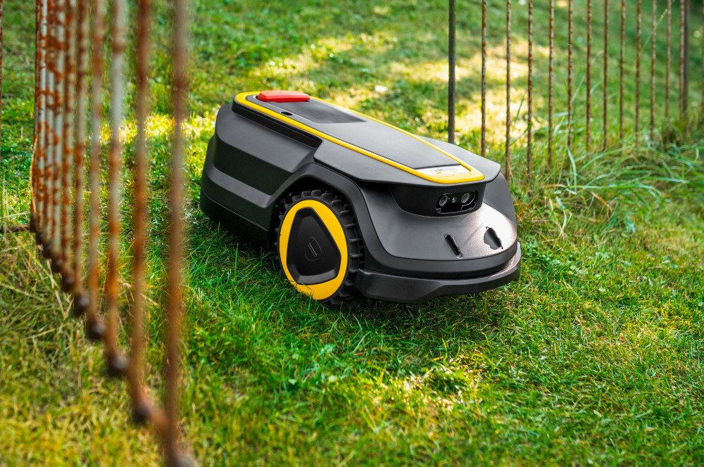 Robot koszący o czarno-żółtej kolorystyce porusza się po trawie obok metalowego ogrodzenia. Urządzenie ma kompaktowy kształt i widoczne koła z żółtymi akcentami.