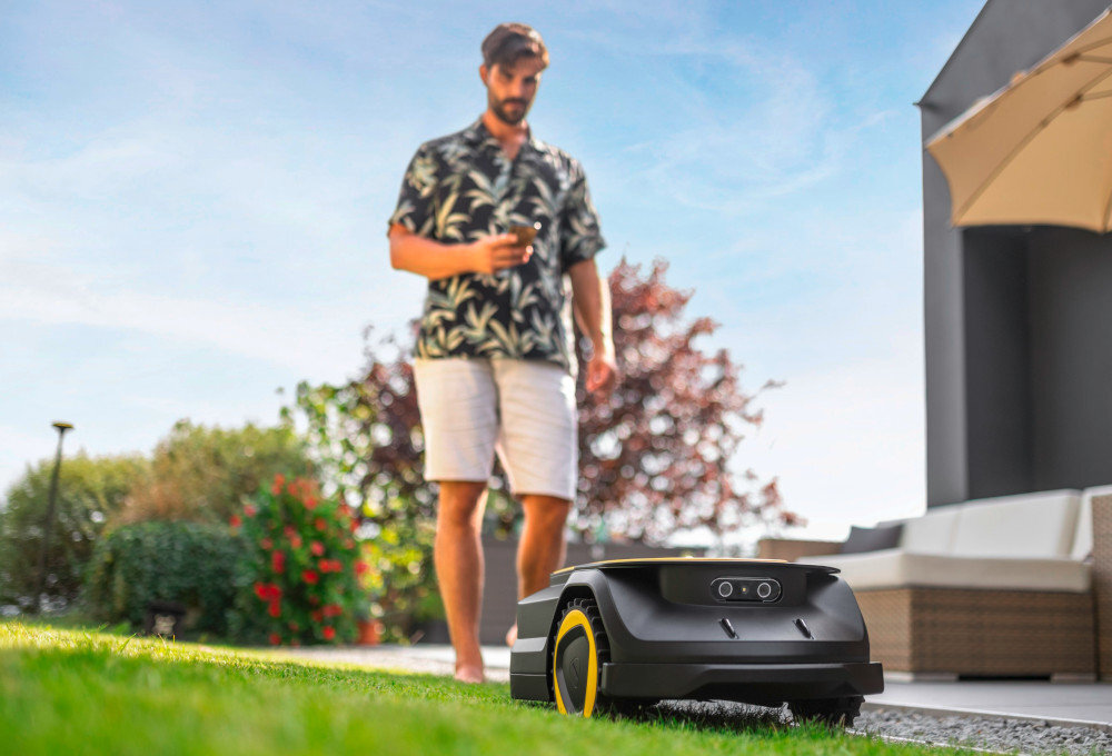 Na trawniku znajduje się robot koszący z żółtymi kołami. W tle stoi osoba trzymająca urządzenie, a obok widoczny jest parasol ogrodowy i meble tarasowe.