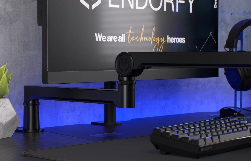 Na biurku znajduje się czarne ramię mikrofonowe zamontowane obok monitora z logo ENDORFY. Na ekranie widoczny jest tekst: \'We are all technology heroes\'.