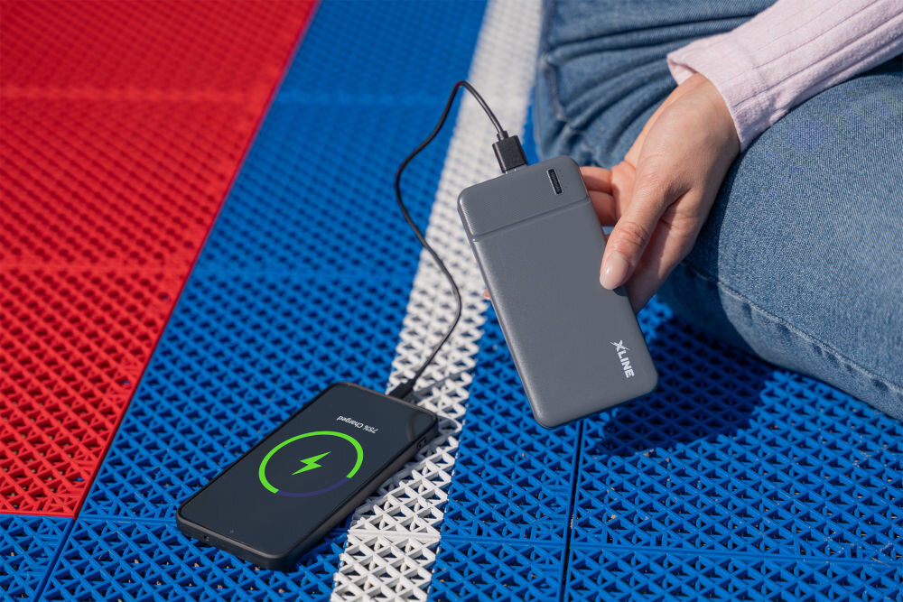 Osoba trzyma szary powerbank z logo XLINE, który jest podłączony do smartfona leżącego na kolorowej powierzchni. Na ekranie telefonu widoczny jest symbol ładowania i napis \'media expert\'.