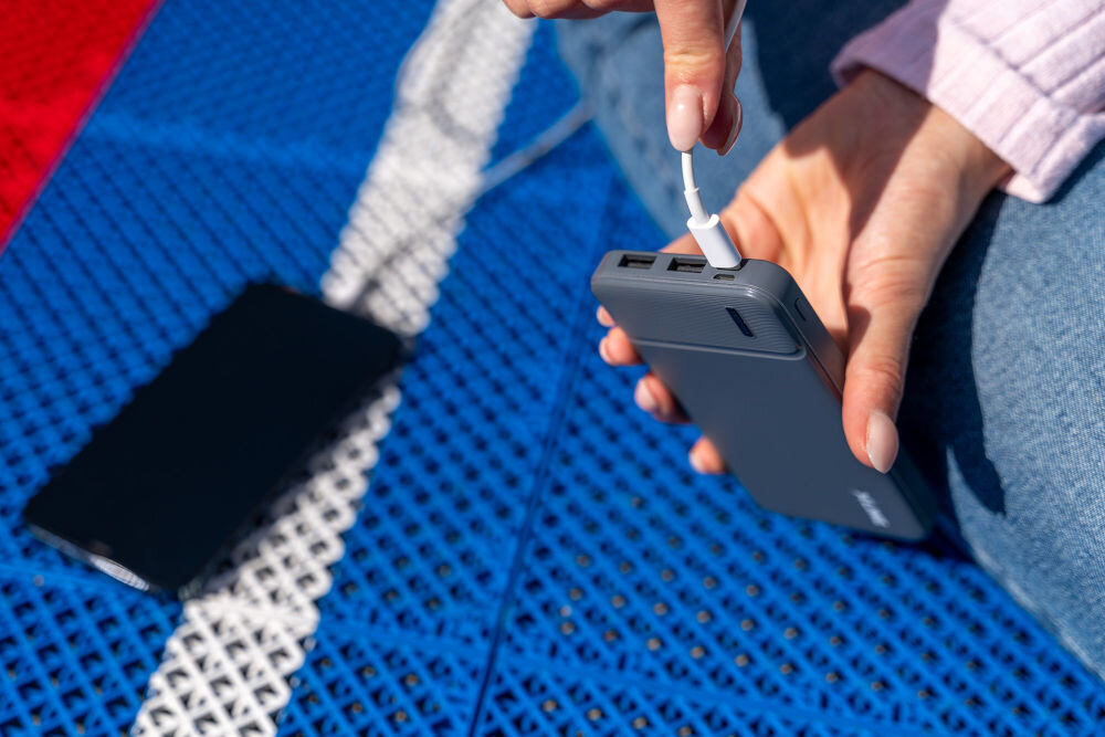 Osoba trzyma szary powerbank, do którego podłączony jest kabel USB. W tle widać telefon leżący na niebiesko-czerwonej powierzchni.
