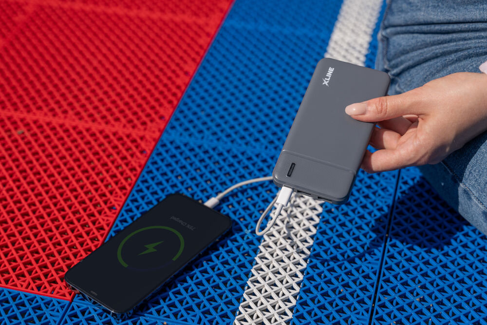Osoba trzyma szary powerbank z logo \'XLINE\', który ładuje telefon leżący na kolorowej powierzchni z czerwonym, niebieskim i białym wzorem.