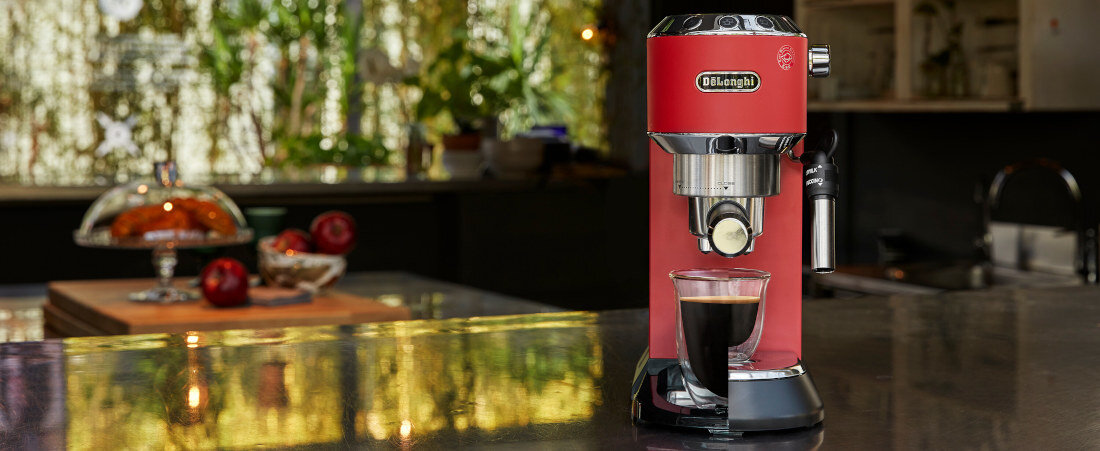 Czerwony ekspres do kawy z logo \'DeLonghi\' stoi na blacie kuchennym, obok szklanki z kawą. W tle widać rozmytą kuchnię z roślinami i owocami na stole.