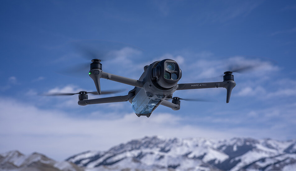 Dron z czterema wirnikami unosi się w powietrzu na tle ośnieżonych gór i niebieskiego nieba. Na dronie widoczny jest napis \'Mavic 4 Pro\'.
