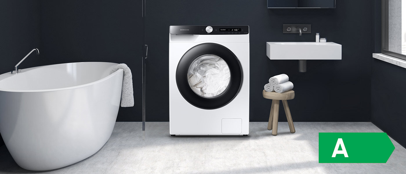 Zdjęcie pralki Samsung wraz z ikoną oznaczającą najwyższą klasę energetyczną W łazience znajduje się biała pralka z czarnym panelem, obok wolnostojąca wanna i umywalka. Na podłodze leży zielony prostokąt z literą \'A\'.