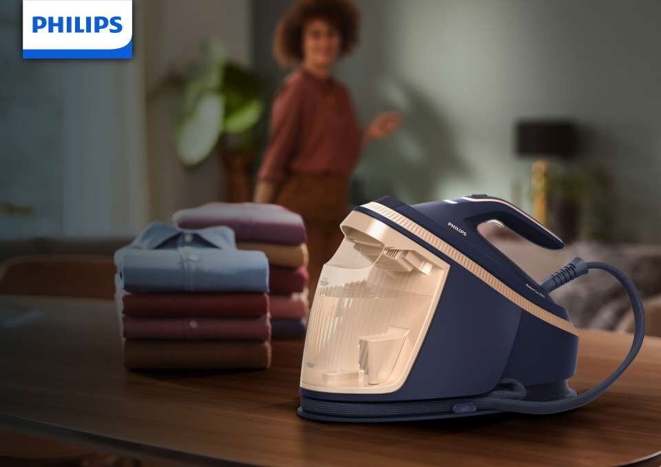 Na drewnianym stole znajduje się generator pary w kolorze granatowym z logo PHILIPS. Obok leży stos złożonych koszul, a w tle widoczna jest osoba w pomieszczeniu.
