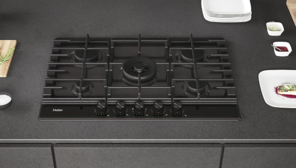 Czarna płyta gazowa z pięcioma palnikami i pokrętłami do regulacji, umieszczona na ciemnym blacie kuchennym. Widoczne logo: Haier.