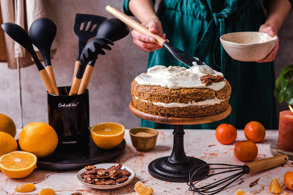 Na stole znajduje się tort na paterze, obok którego leżą pomarańcze, orzechy i trzepaczka. W tle widoczny jest zestaw przyborów kuchennych w czarnym pojemniku z napisem \'Cook with love\'.