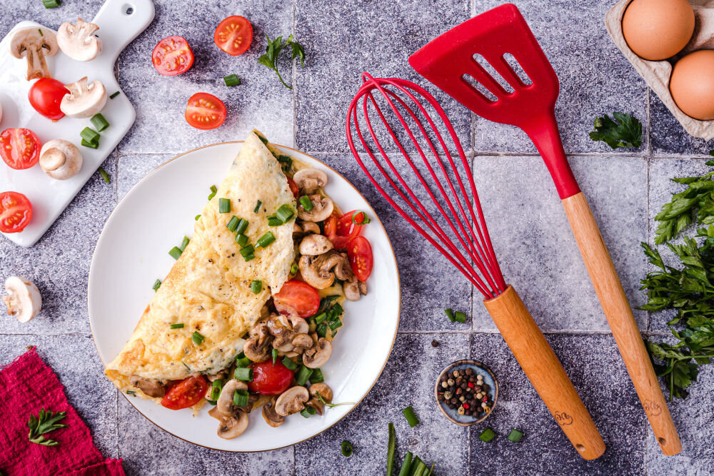 Na talerzu znajduje się omlet z pieczarkami, pomidorami i szczypiorkiem. Obok leżą czerwone przybory kuchenne oraz składniki takie jak jajka, pieczarki i pomidory koktajlowe.
