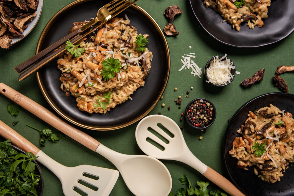 Na zielonym tle znajdują się talerze z risotto, udekorowane natką pietruszki i startym serem. Obok leżą drewniane przybory kuchenne oraz miseczki z przyprawami i suszonymi grzybami.
