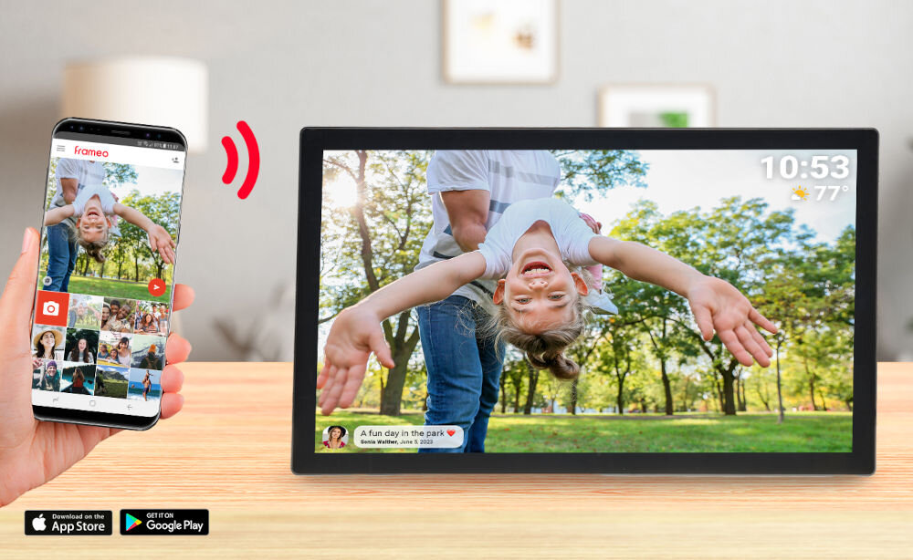 Na stole stoi czarna ramka cyfrowa wyświetlająca zdjęcie dziecka trzymanego do góry nogami w parku. Obok znajduje się smartfon z aplikacją do zarządzania zdjęciami, a na ekranie ramki widoczny jest tekst: \'A fun day in the park ❤️ Bruno Walker - June 5, 2021\'.