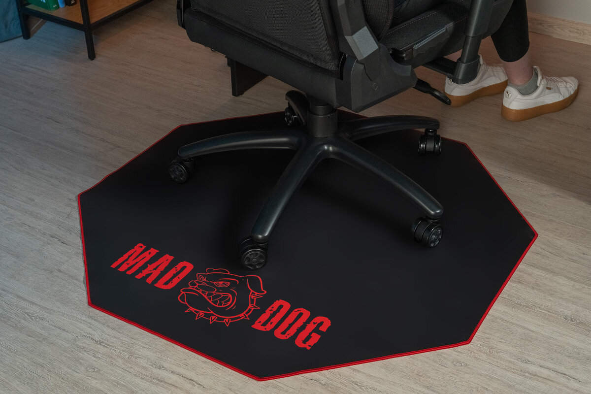 Czarna mata pod fotel gamingowy z czerwonym napisem \'MAD DOG\' i grafiką psa, umieszczona na jasnej podłodze. Na macie stoi czarny fotel biurowy na kółkach, a obok widać nogi osoby w białych butach.