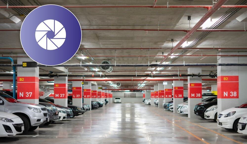 Podziemny parking z zaparkowanymi samochodami po obu stronach. Na kolumnach widoczne są oznaczenia B2, N 37, N 38.