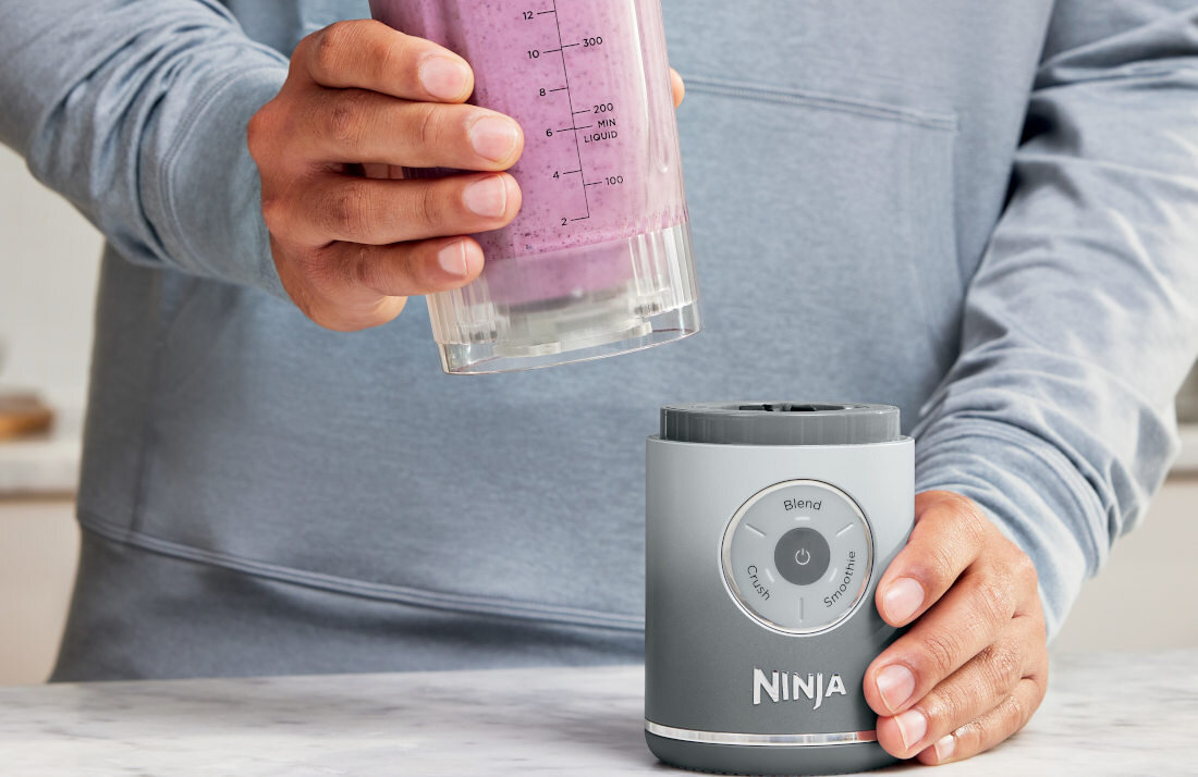 Osoba trzyma kubek z fioletowym napojem, a obok znajduje się blender z widocznym logo \'Ninja\'.