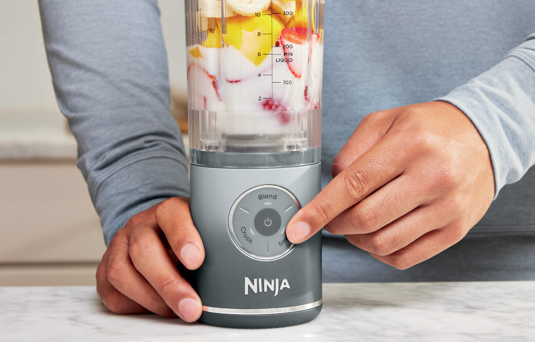 Osoba obsługuje blender z logo \'NINJA\', w którym znajduje się mieszanka owoców. Na blenderze widoczne są przyciski \'Blend\' i \'Crush\'.