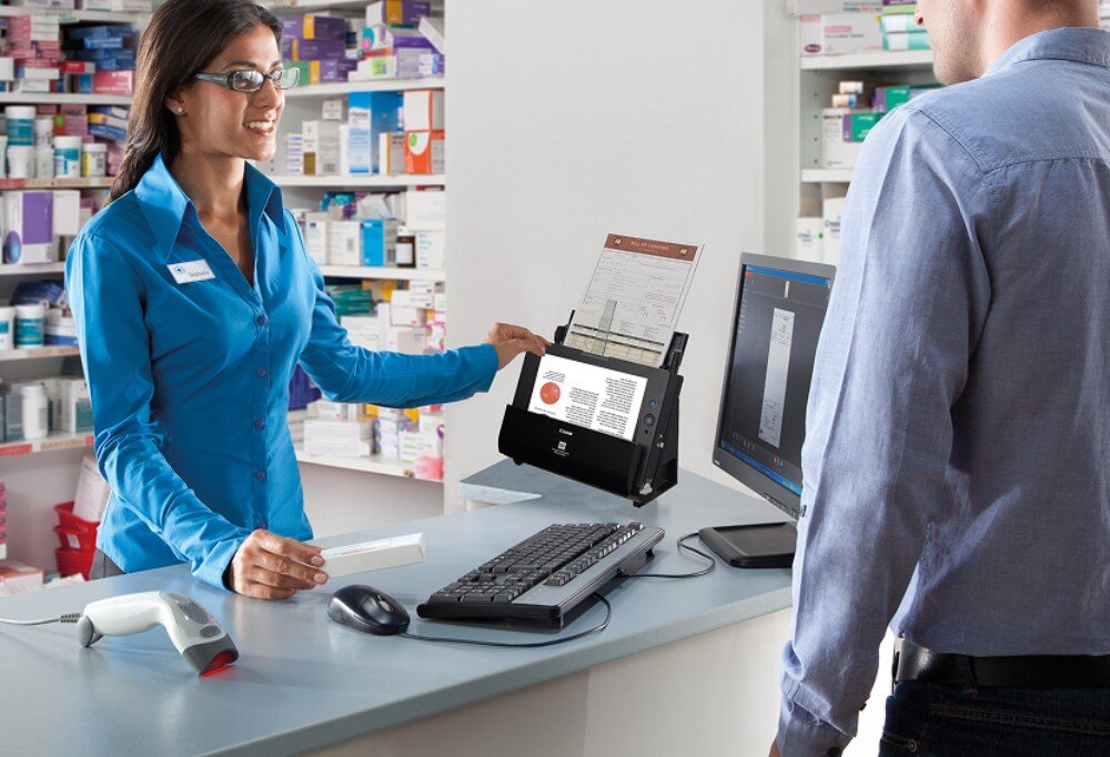 Skaner CANON imageFORMULA DR-C225II Farmaceutka w niebieskiej koszuli i okularach stoi za ladą apteczną, uśmiechając się do klienta w szarej koszuli. Na blacie obok klawiatury i myszki znajduje się pionowy czarny skaner, opis