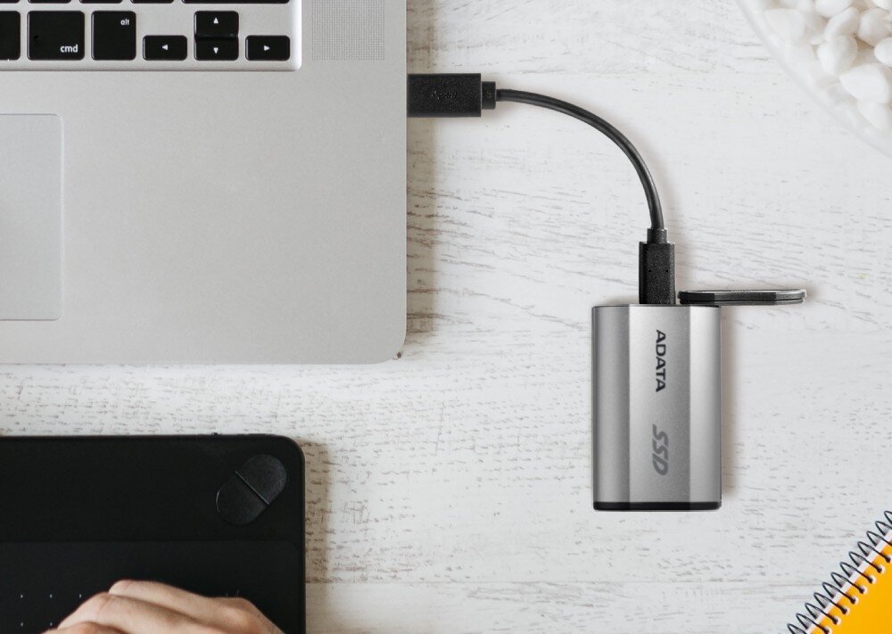 Na białym stole znajduje się srebrny dysk SSD marki ADATA podłączony do laptopa za pomocą kabla USB. Obok widoczna jest część tabletu oraz miska z białymi przedmiotami.