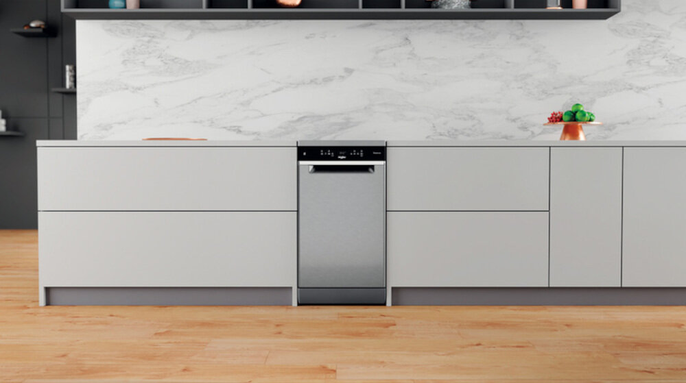 Zmywarka w kolorze inox wbudowana w nowoczesną kuchenną zabudowę z białymi frontami. Na blacie widoczne są owoce na paterze oraz deska do krojenia.