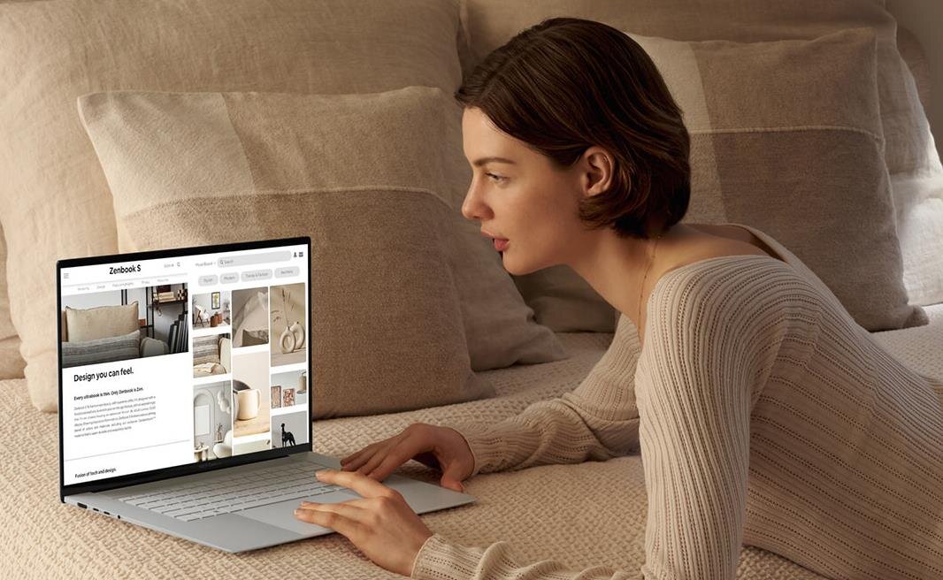 Osoba leży na łóżku i używa laptopa, na ekranie którego widoczna jest strona internetowa z nagłówkiem \'Zenbook S\'.