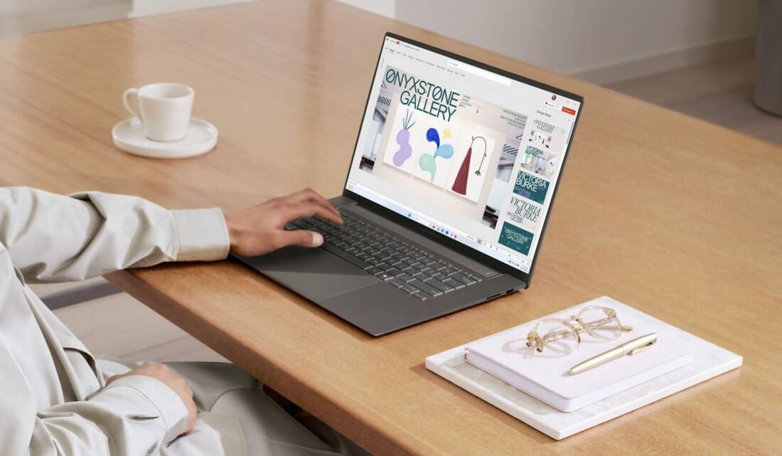 Osoba korzysta z laptopa, na ekranie którego widoczna jest strona z napisem \'ONYXSTONE GALLERY\'. Obok laptopa na stole leży filiżanka oraz notes z okularami i długopisem.