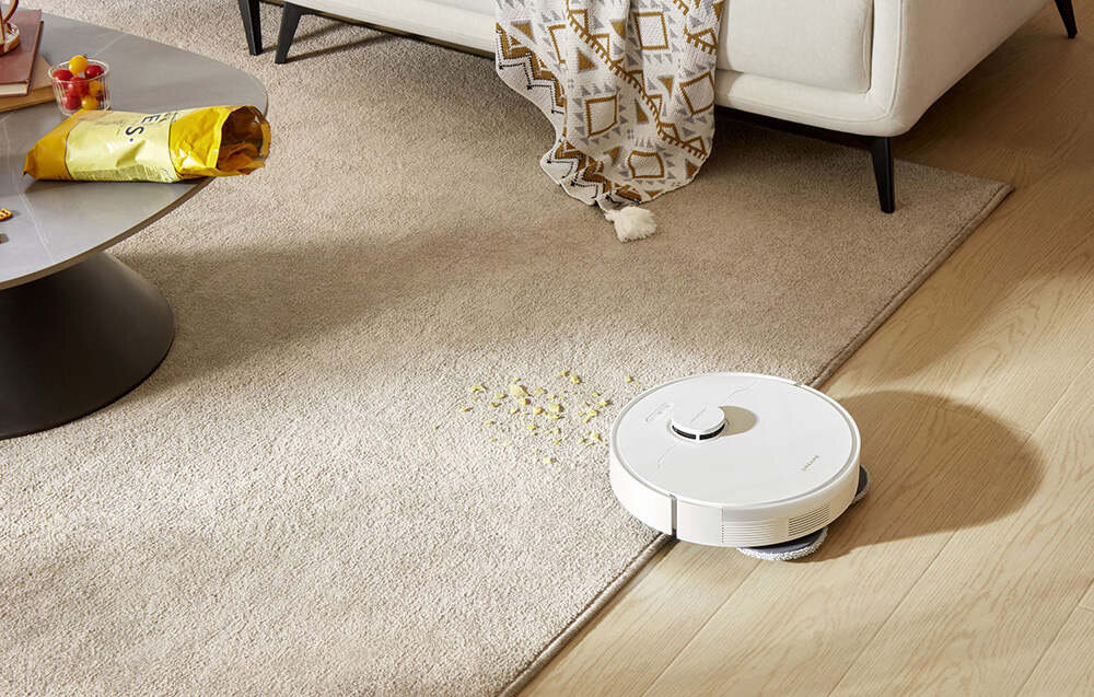 Na podłodze widać biały robot sprzątający, który znajduje się na granicy dywanu i drewnianej podłogi. Obok robota leżą rozsypane drobinki, a w tle stoi stolik z otwartą paczką chipsów.