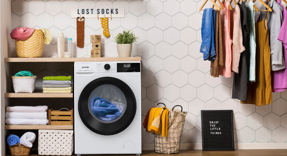 Pralka z logo Gorenje stoi w pomieszczeniu z półkami pełnymi ręczników i koszyków. Na ścianie wisi tabliczka z napisem \'LOST SOCKS\', a obok pralki znajduje się kosz z praniem i tabliczka z napisem \'ENJOY THE LITTLE THINGS\'.