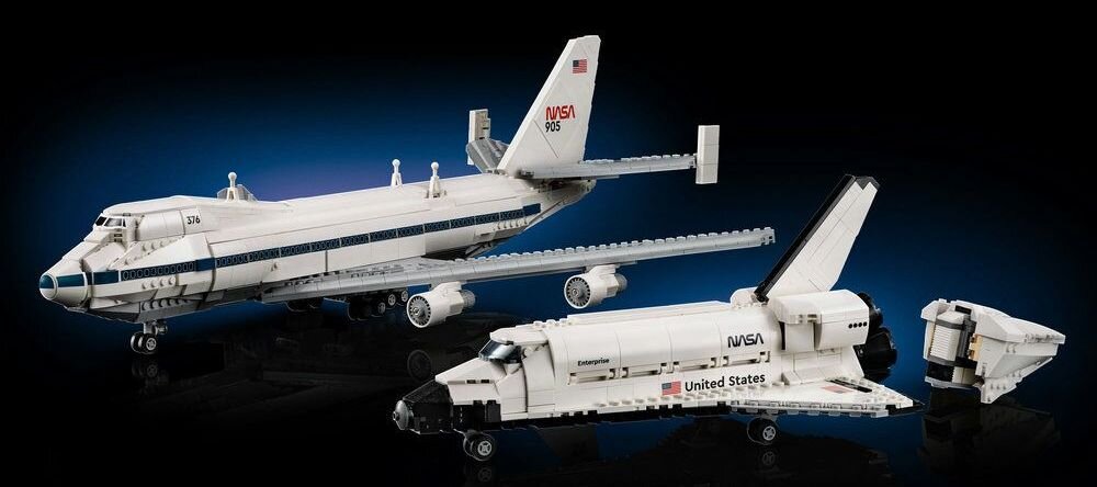 Na obrazie widoczne są dwa modele pojazdów kosmicznych z logo NASA, w tym samolot transportowy i prom kosmiczny. Modele są szczegółowo wykonane i ustawione na ciemnym tle.