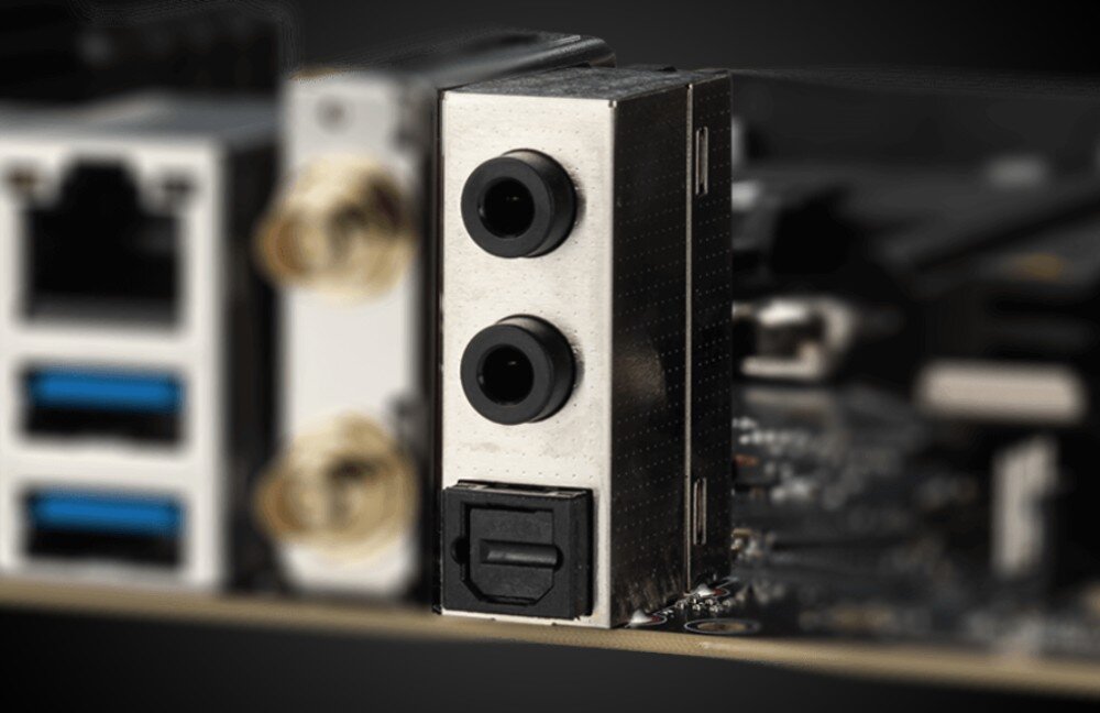 Zbliżenie na panel złącz płyty głównej, widoczne są dwa porty USB, dwa gniazda audio oraz port optyczny. W tle widać fragmenty innych złącz i elementów płyty głównej.