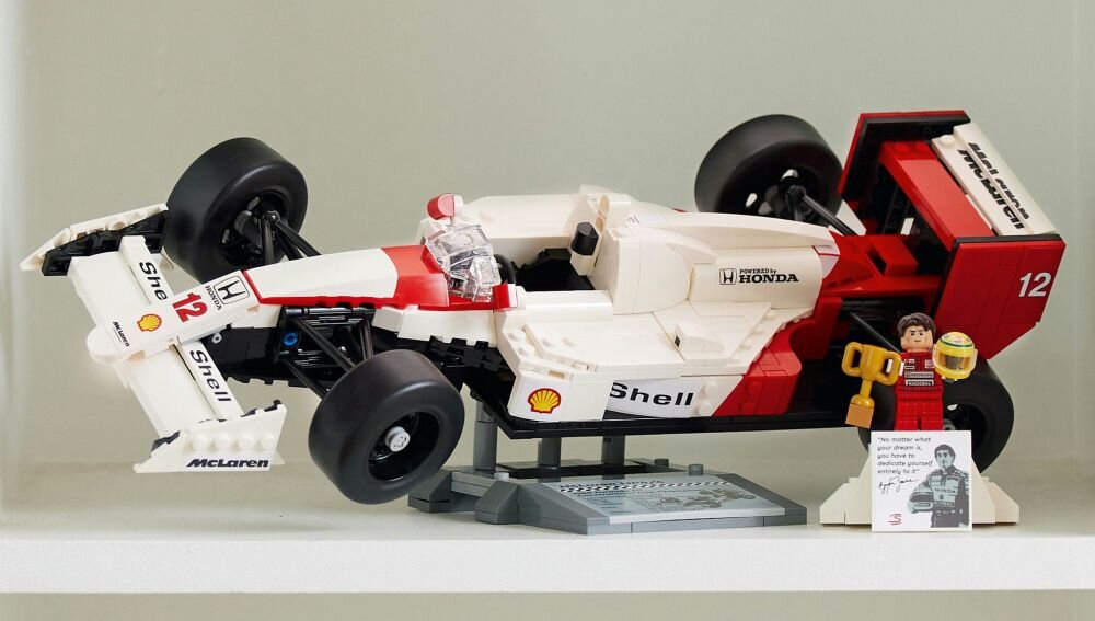 Model samochodu wyścigowego z klocków LEGO w kolorach białym, czerwonym i czarnym z widocznymi logotypami Shell, Honda i McLaren. Obok modelu stoi figurka z pucharem oraz tabliczka z czarno-białym zdjęciem i tekstem.