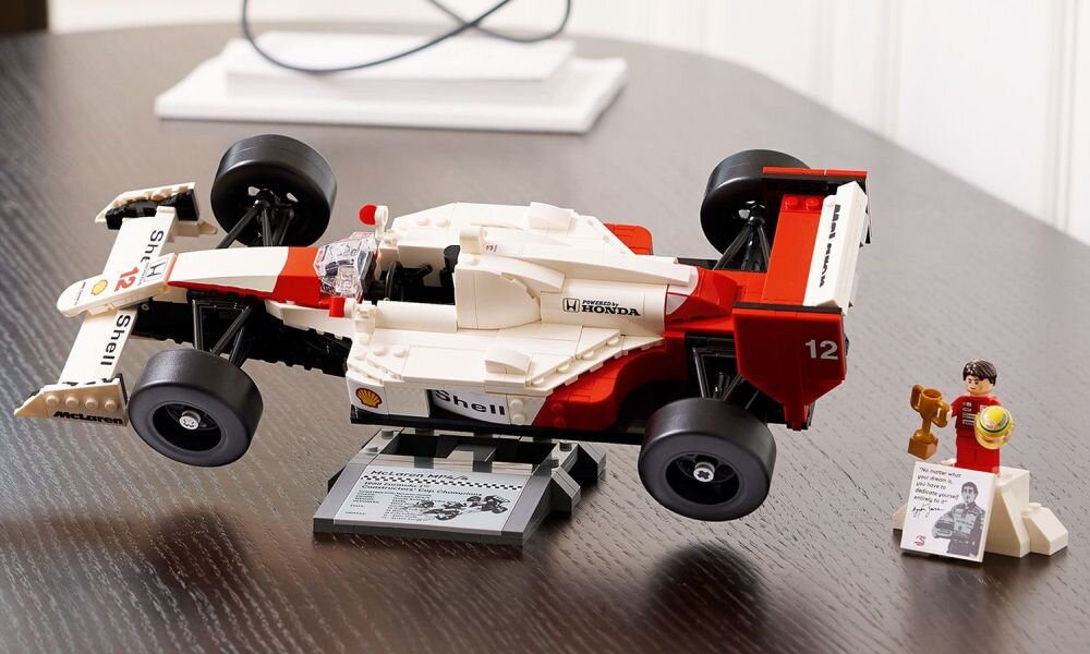 Model samochodu wyścigowego z klocków LEGO w kolorach białym i czerwonym z widocznymi logotypami \'Shell\' i \'Honda\'. Obok znajduje się figurka z pucharem i tabliczką z czarno-białym zdjęciem.