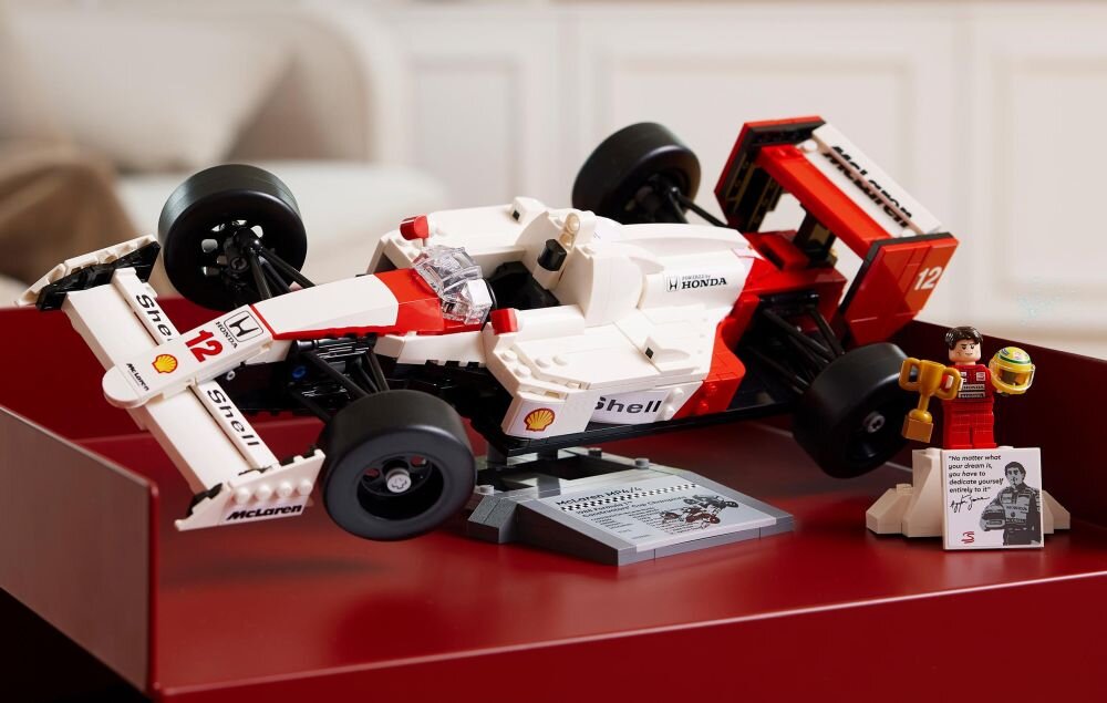 Model samochodu wyścigowego z klocków LEGO w kolorach białym, czerwonym i czarnym, z widocznymi logotypami Shell, Honda i McLaren. Obok znajduje się figurka z kaskiem oraz tabliczka z napisem \'The greatest driver of all time\' i podpisem Ayrton Senna.